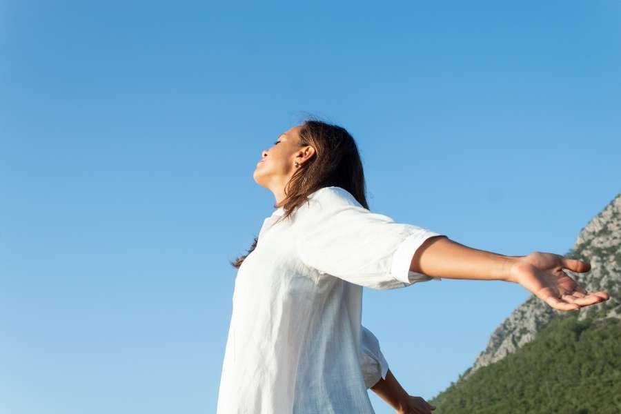 A woman in a white shirt standing outdoors with arms wide open, experiencing somatic healing.