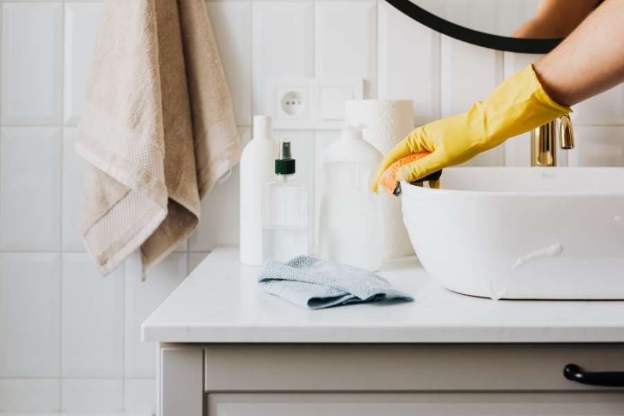 A person wearing a yellow cleaning glove cleans a modern white bathroom sink basin.